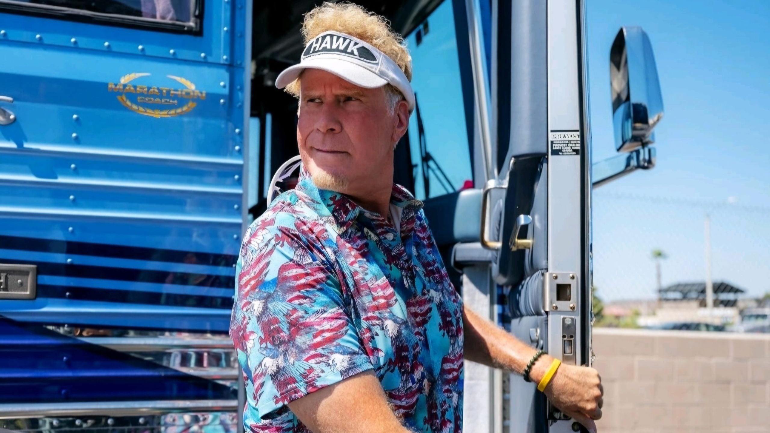 A man in a colorful shirt and visor stands at the open door of a blue bus, looking to the side on a sunny day.