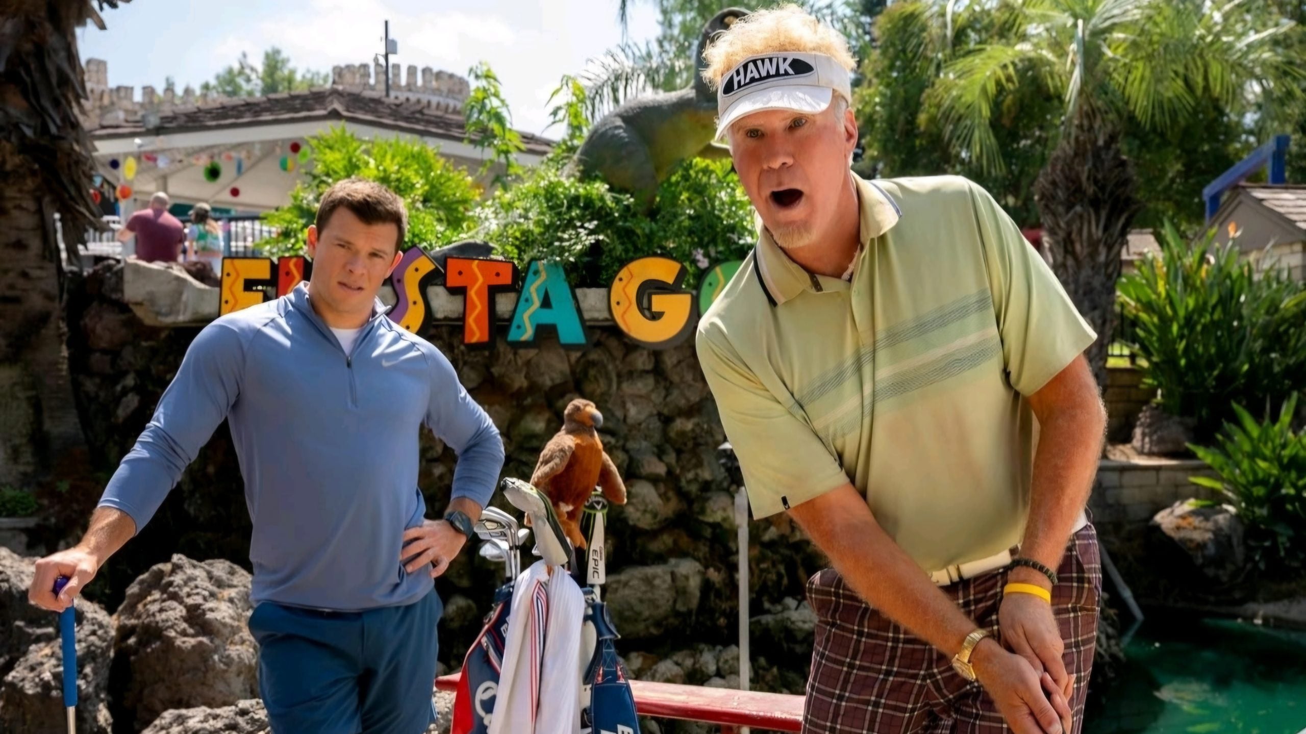 Two men are playing mini golf outdoors; one is about to take a shot while the other stands nearby with a bird perched on their golf bag. Colorful "FIESTA" letters are in the background.