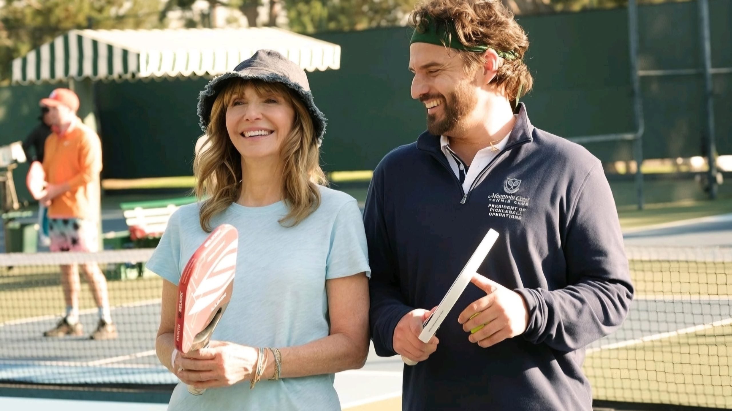 Two people smiling and holding pickleball paddles stand on a court, with another person in the background, all dressed in casual sportswear.