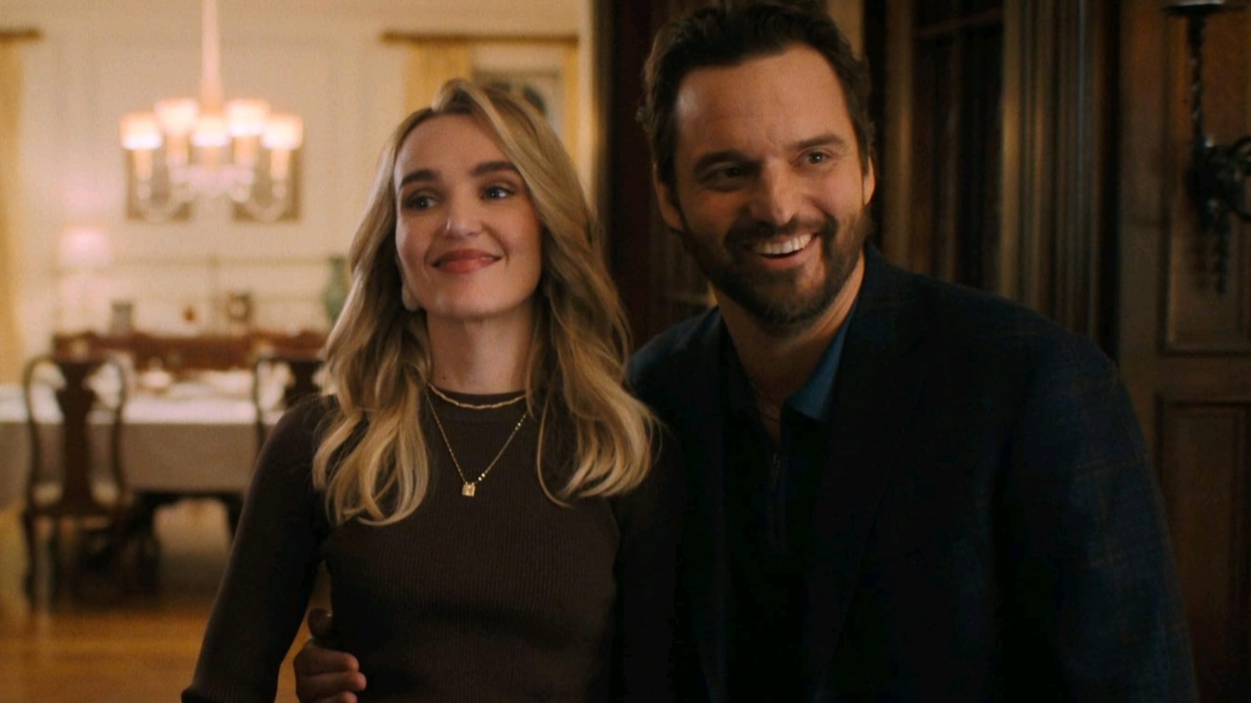 A man and a woman stand close together indoors, smiling and looking off to the side. The background features warm lighting and classic decor.