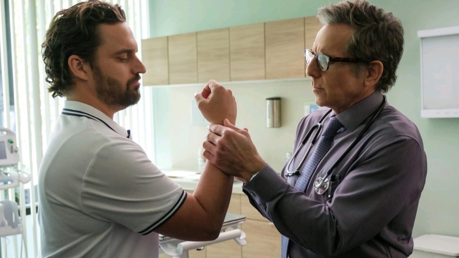‘The Dink’ Review: Very Funny And Irreverent A doctor examines a man's wrist in a medical office. The man wears a white polo shirt, and the doctor is dressed in a shirt with a stethoscope. Cabinets and medical equipment are visible.