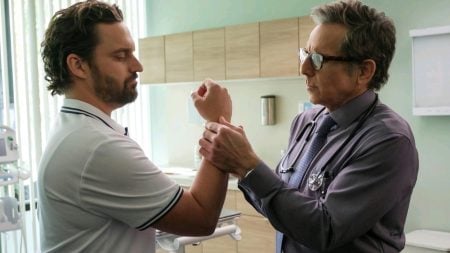 ‘The Dink’ Review: Very Funny And Irreverent A doctor examines a man's wrist in a medical office. The man wears a white polo shirt, and the doctor is dressed in a shirt with a stethoscope. Cabinets and medical equipment are visible.