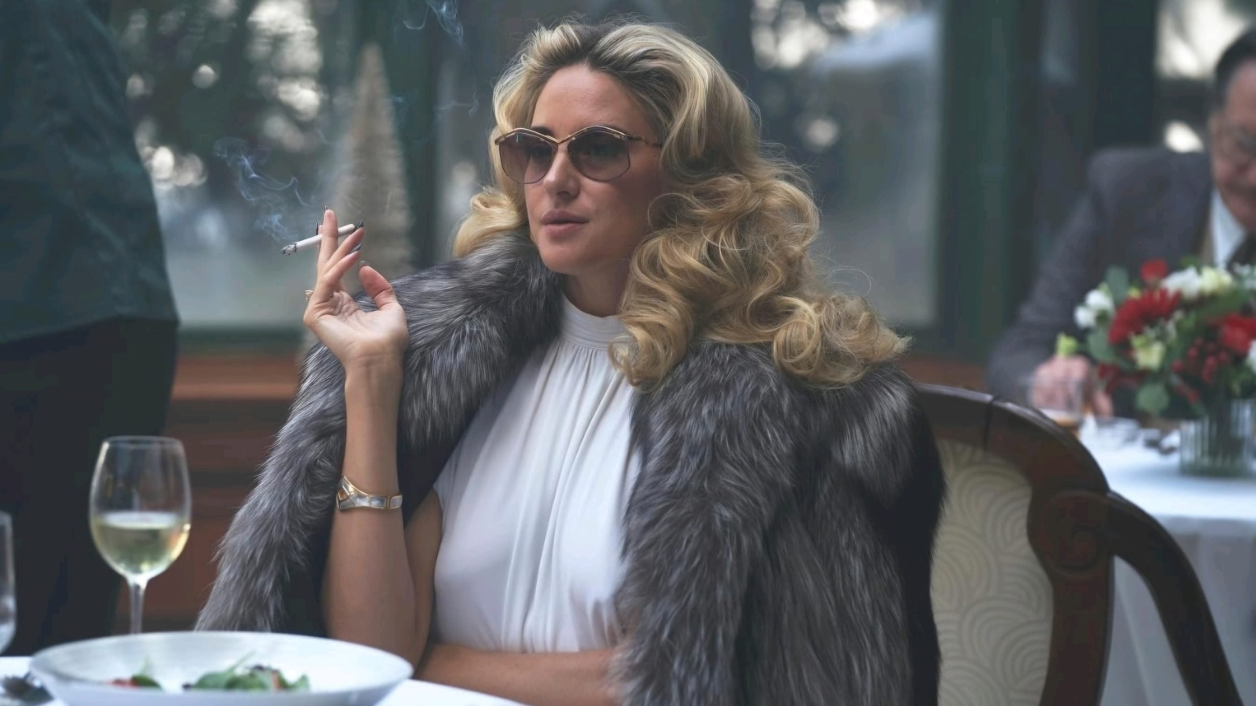 Woman with wavy blond hair, wearing sunglasses and a fur coat, sits at a table with a salad and wine, holding a cigarette.