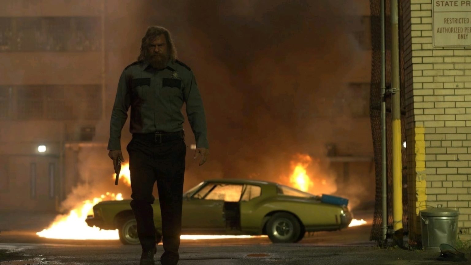‘Motor City’ Review: Alan Ritchson Lets His Fists Do The Talking In A Scintillating Silent-Era Throwback A man in a uniform holding a gun walks away from a burning car at night, with flames and smoke in the background.