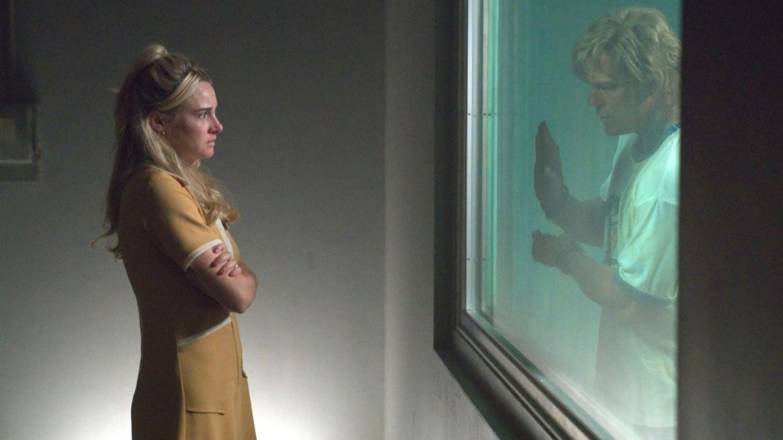 A woman in a yellow dress stands with arms crossed, facing a man behind glass who presses his hand to the window.