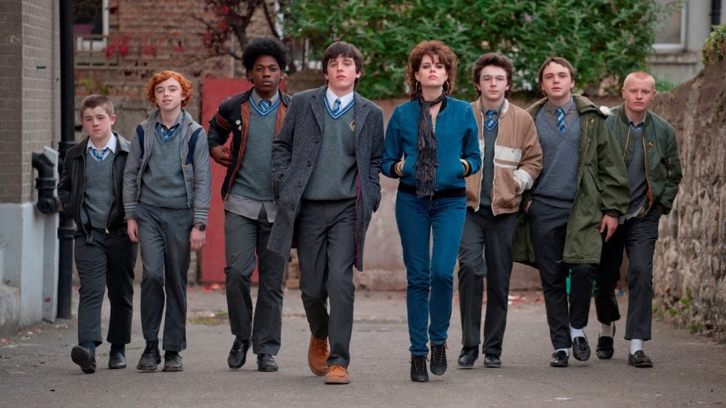 A group of teenagers in school uniforms and one girl in casual clothes walk side by side down a street, looking ahead confidently.