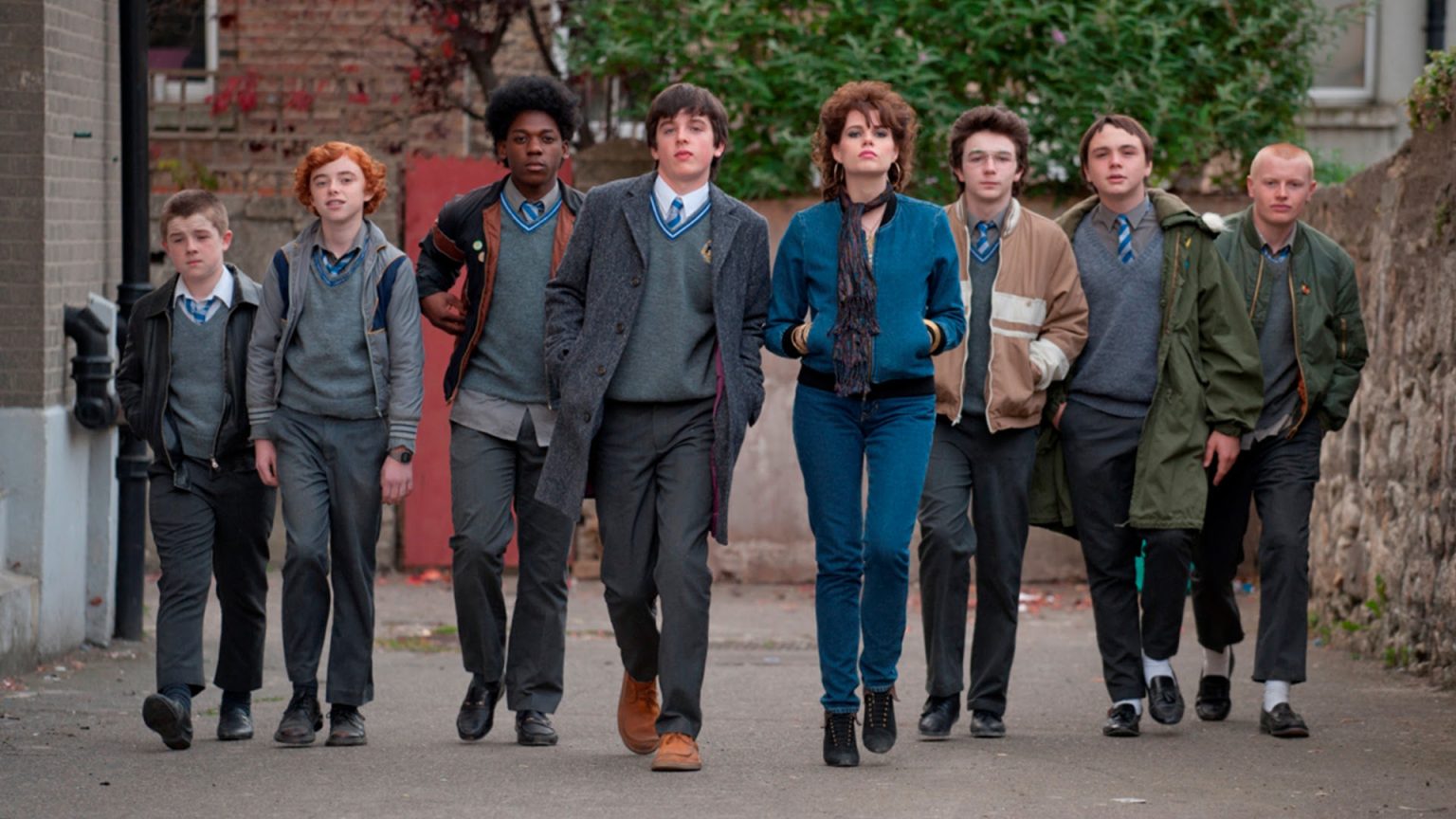 A group of teenagers in school uniforms and one girl in casual clothes walk side by side down a street, looking ahead confidently.