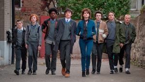 A group of teenagers in school uniforms and one girl in casual clothes walk side by side down a street, looking ahead confidently.