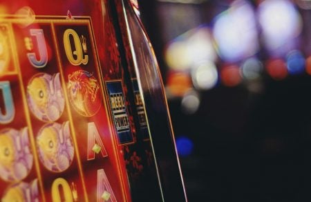 Close-up of a brightly lit casino slot machine with colorful symbols and a blurred background of other machines and lights.