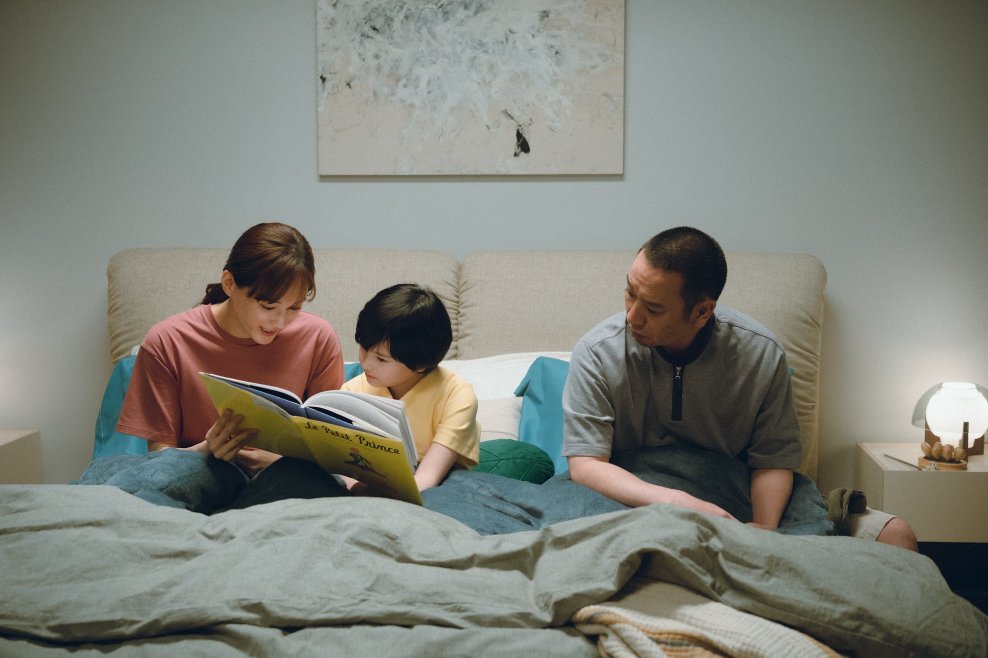 A woman reads a large picture book to a child while a man sits beside them on a bed in a softly lit bedroom.