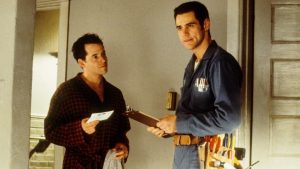 Two men stand indoors; one wears a robe and holds an envelope, while the other wears a blue work uniform with tools and holds a clipboard.