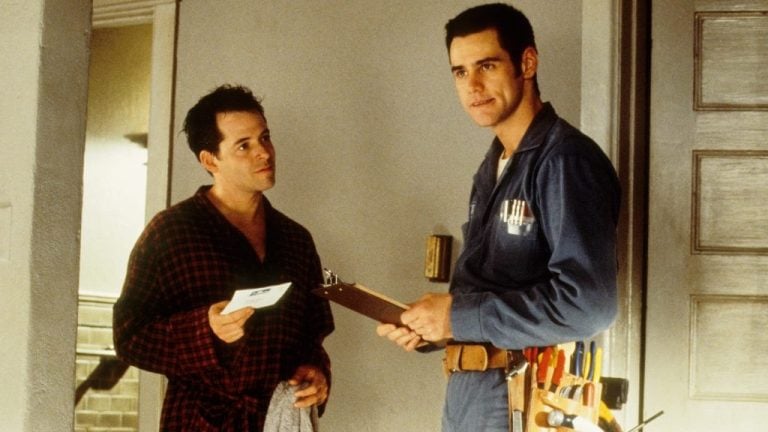 Sony To Celebrate The 30th Anniversary Of ‘The Cable Guy’ With 4K UHD Blu-Ray Debut This October Two men stand indoors; one wears a robe and holds an envelope, while the other wears a blue work uniform with tools and holds a clipboard.