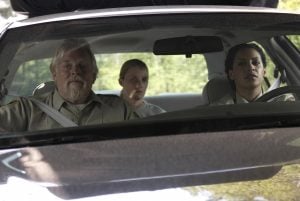 Three people sit in a car; two wear police uniforms in the front seats, and one person sits in the back. Sunlight and greenery are visible through the windows.