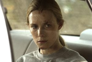 A woman with visible scratches and bruises on her face sits in the backseat of a car, looking serious and pensive.