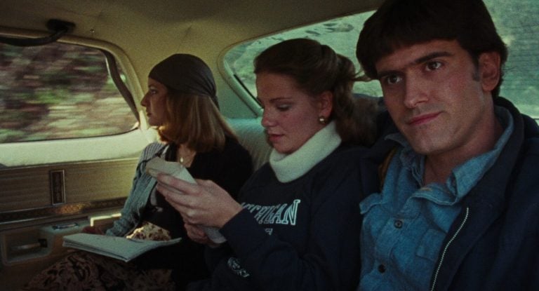 ‘The Evil Dead’ (1981) 4K UHD Blu-Ray SteelBook Review – Sam Raimi’s Gross-Out Debut Is A Classic Three people sit in the backseat of a car; the woman on the left reads, the woman in the middle looks at a piece of paper, and the man on the right looks ahead.