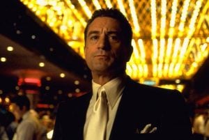 A man in a dark suit and white tie stands in a casino with bright, golden lights above him.