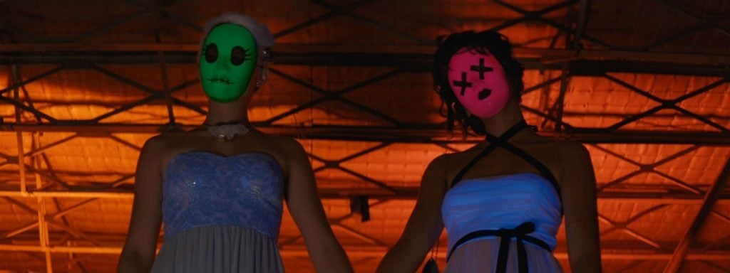 Two people in strapless dresses wear colorful, expressionless masks and stand under an orange-lit, industrial ceiling, holding hands.