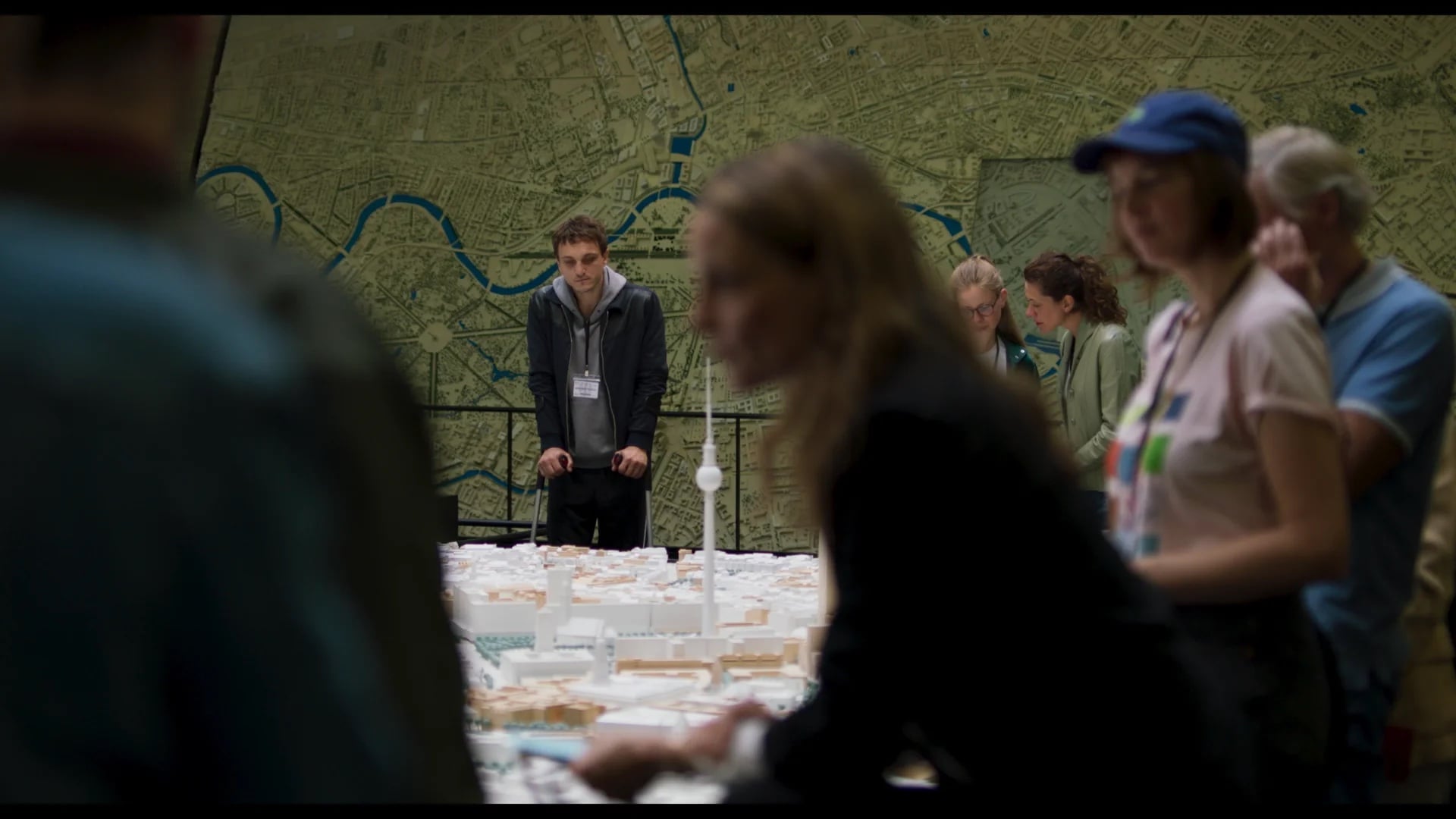 People observe a large city model on a table with a detailed map on the wall behind them; one man stands out in focus at the back, while others are blurred in the foreground.