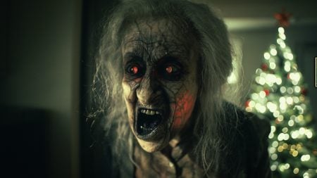 A person in creepy makeup with red eyes and fangs stands in a dim room; a lit Christmas tree is visible in the blurred background.