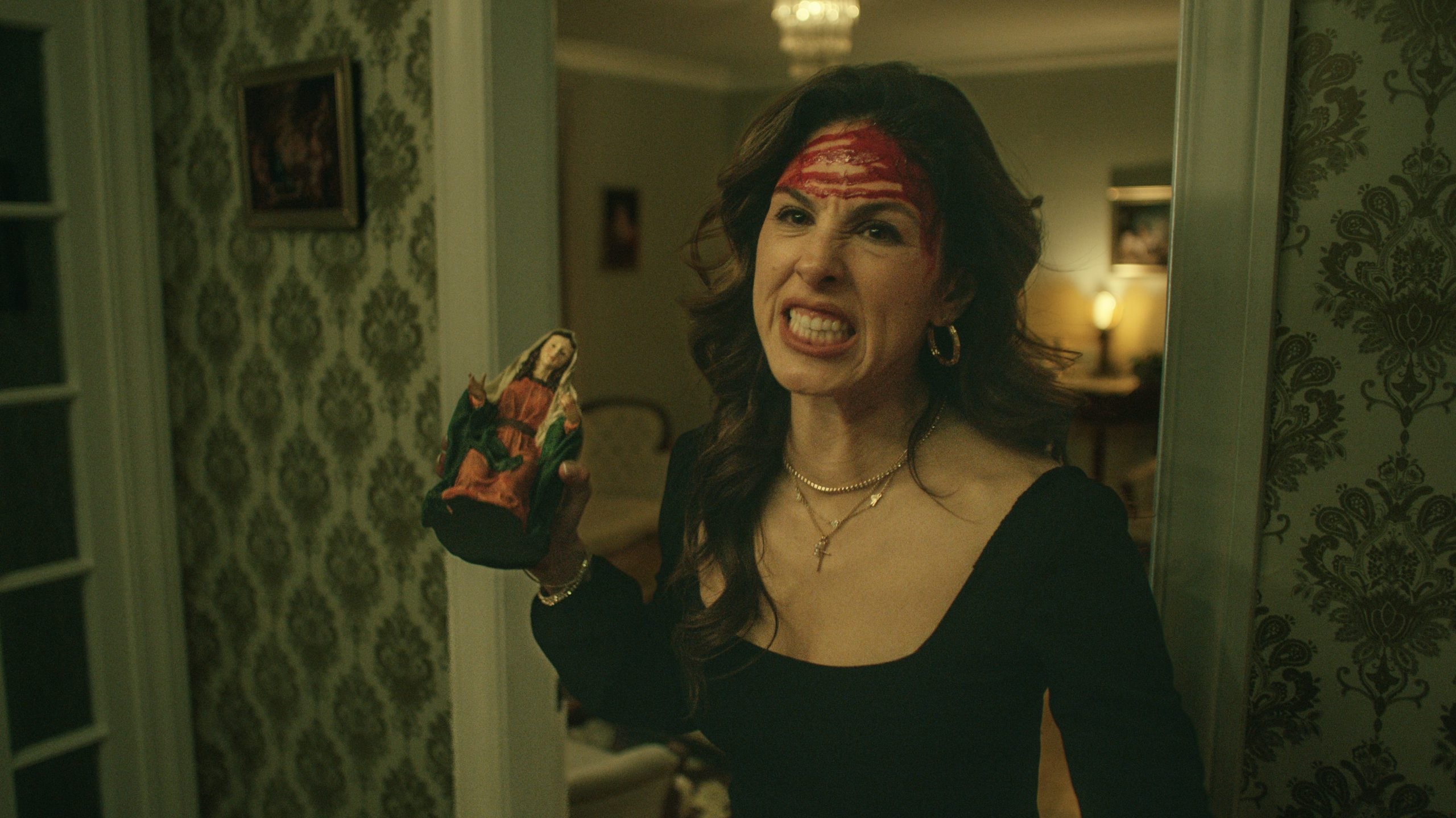 A woman with red markings on her forehead holds a small religious statue and stands in a dimly lit room, showing an intense facial expression.