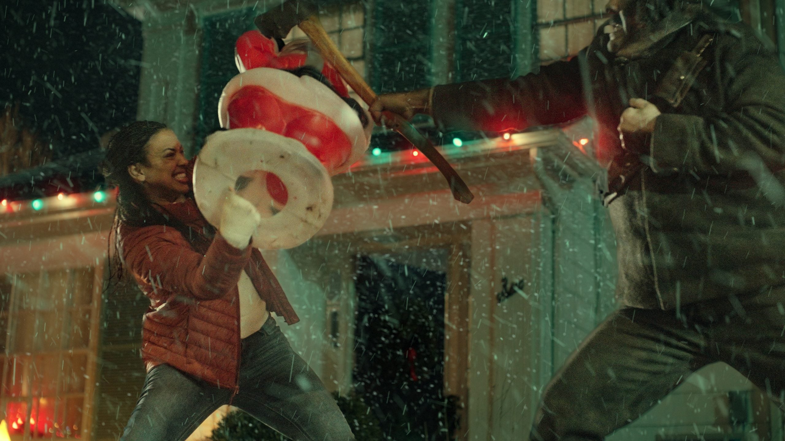 Two people fight outside a house in the snow; one swings an inflatable Santa, the other wields an axe. Holiday lights are visible on the house behind them.