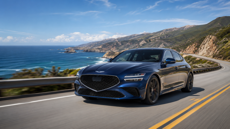 A blue sedan drives along a coastal highway with ocean waves and mountains in the background under a clear blue sky.