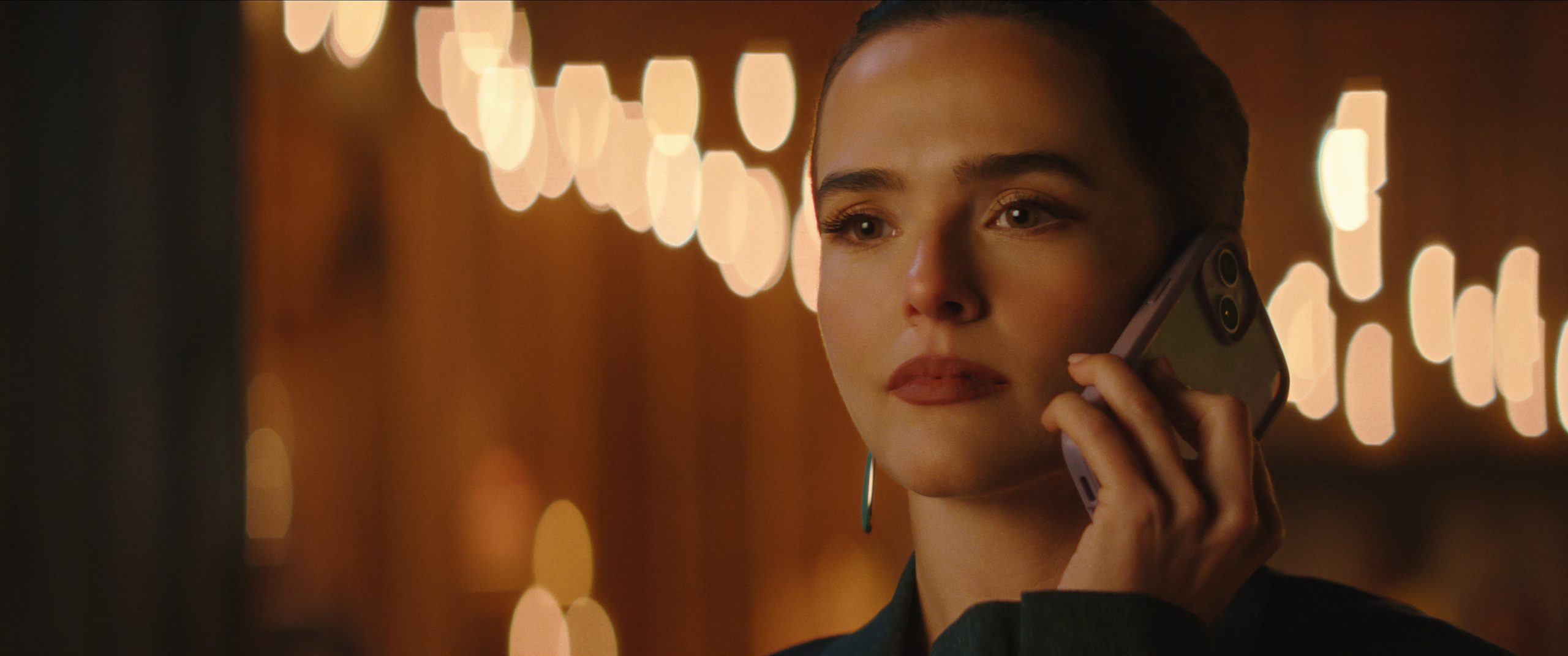 A woman holds a smartphone to her ear and looks ahead with a serious expression. Warm, blurry string lights are visible in the background.