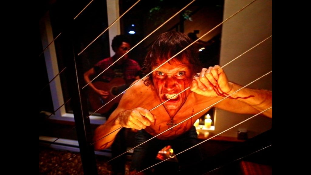 A shirtless person with a fierce expression grips metal wires, with blood on their hand. Candles and another person playing guitar are visible in the background.