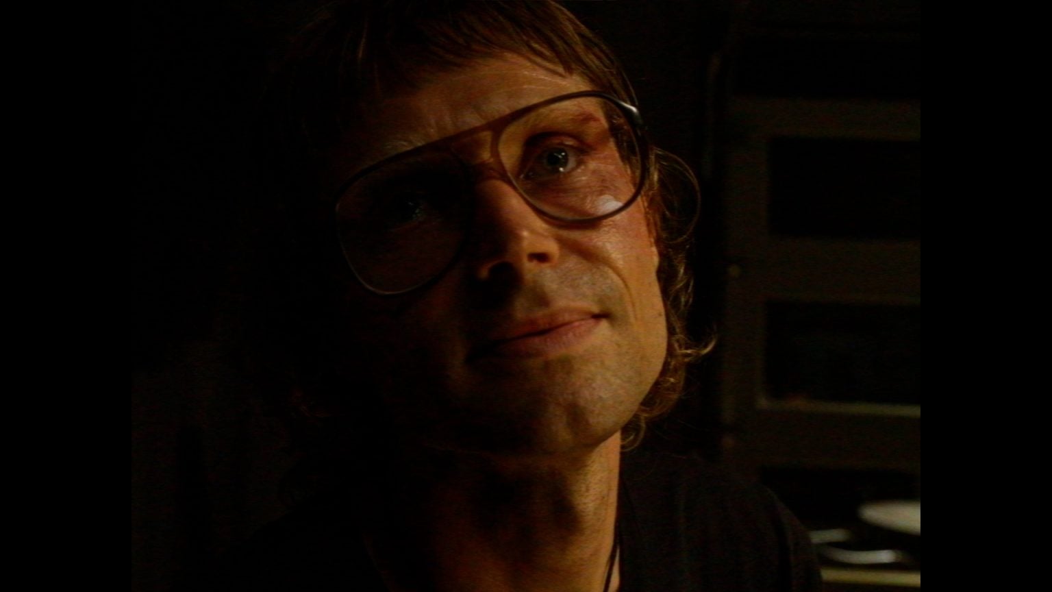 Dark Sky Films Unveils Trailer & September Release Date For Tim Kasher’s ‘Who’s Watching’ A person with light brown hair, wearing large eyeglasses, looks upward in dim lighting.