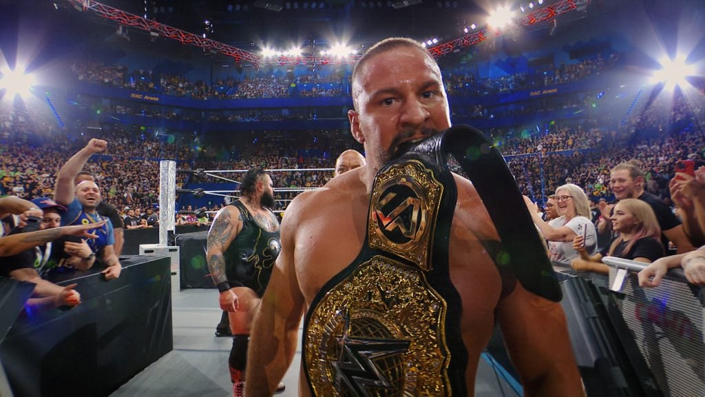 A wrestler holds a championship belt in his mouth while walking through a cheering crowd during a wrestling event.