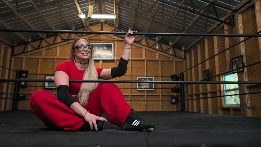 A woman in red pants and a red top sits on the floor of a wrestling ring inside a gym, holding the middle rope and smiling.