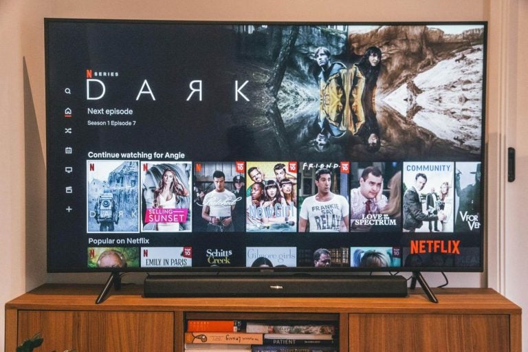 Why Some TV Shows Are Impossible To Watch One Episode At A Time A TV screen displays the Netflix homepage featuring the series “Dark” along with recommendations and trending shows, set on a wooden media console.