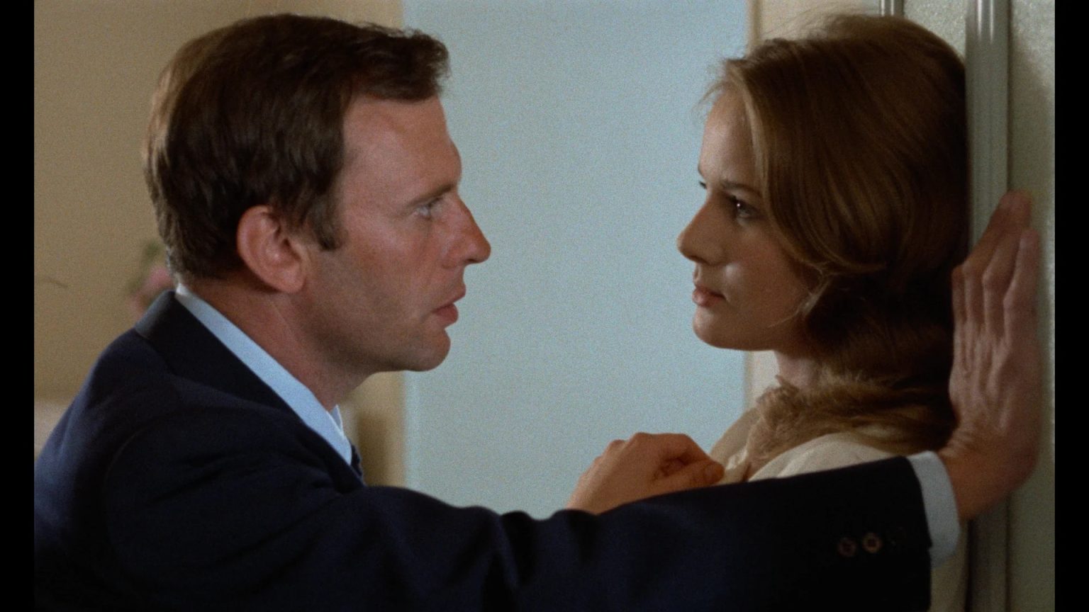 A man in a suit leans against a wall, facing a woman with his arm raised. The woman looks back at him with a neutral expression.