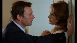 ‘Without Apparent Motive’ Blu-Ray Review – Trintignant Leads Compelling French Crime Thriller A man in a suit leans against a wall, facing a woman with his arm raised. The woman looks back at him with a neutral expression.