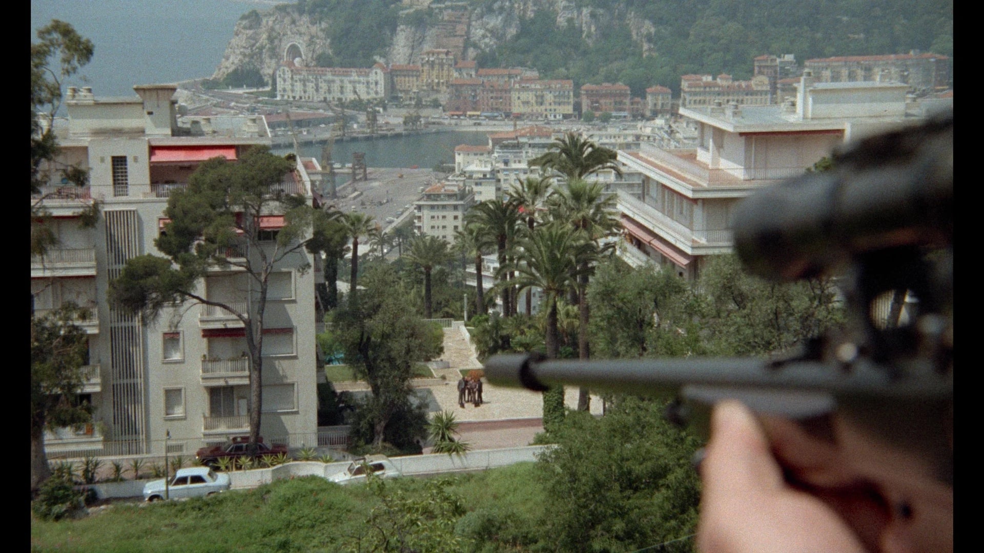 A person aims a scoped rifle from a balcony overlooking a coastal city with buildings, trees, roads, and distant waterfront.