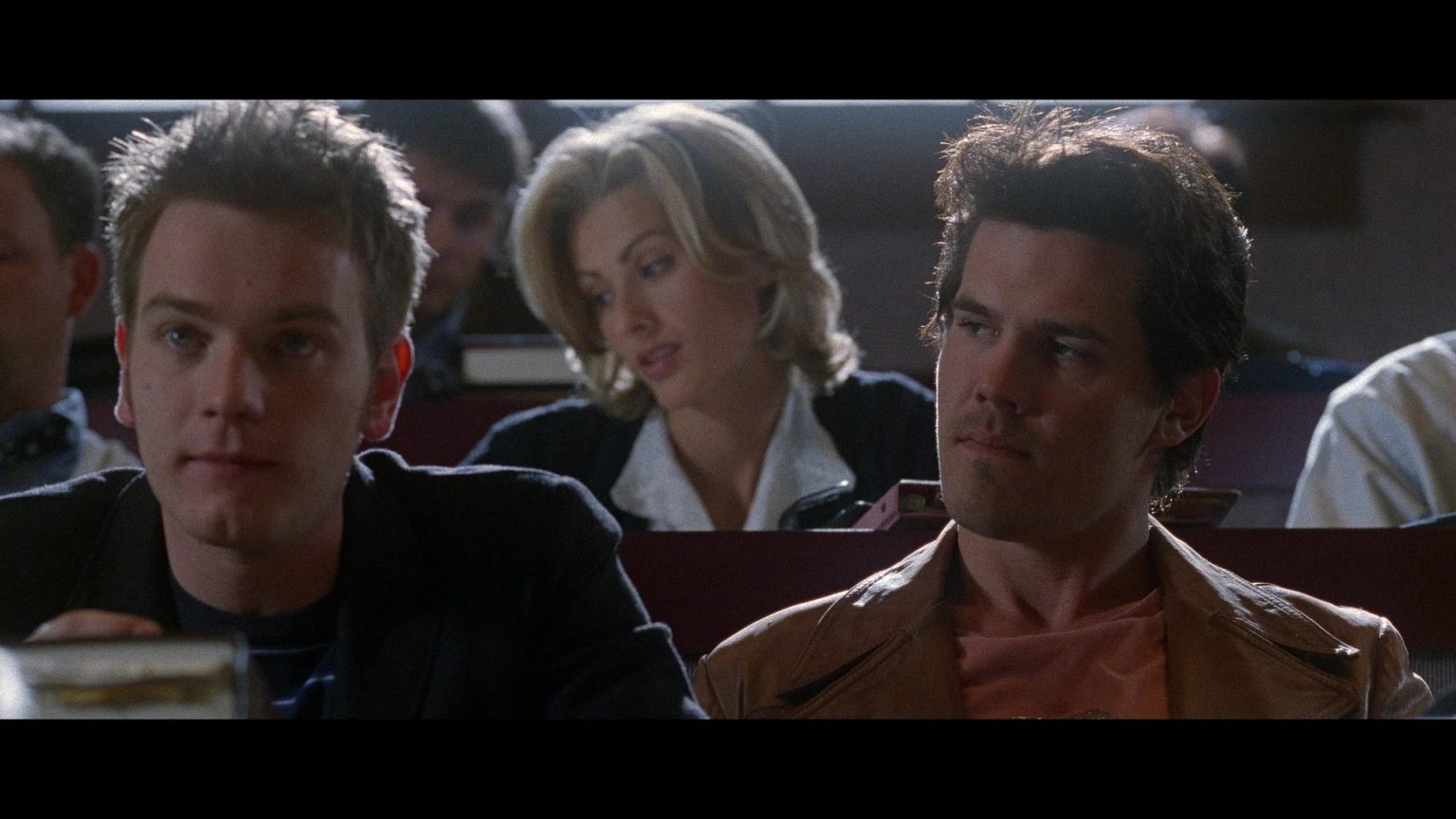 Three people sit in a lecture hall. Two men are seated in the foreground, while a woman sits behind them, looking down. Other students appear out of focus in the background.
