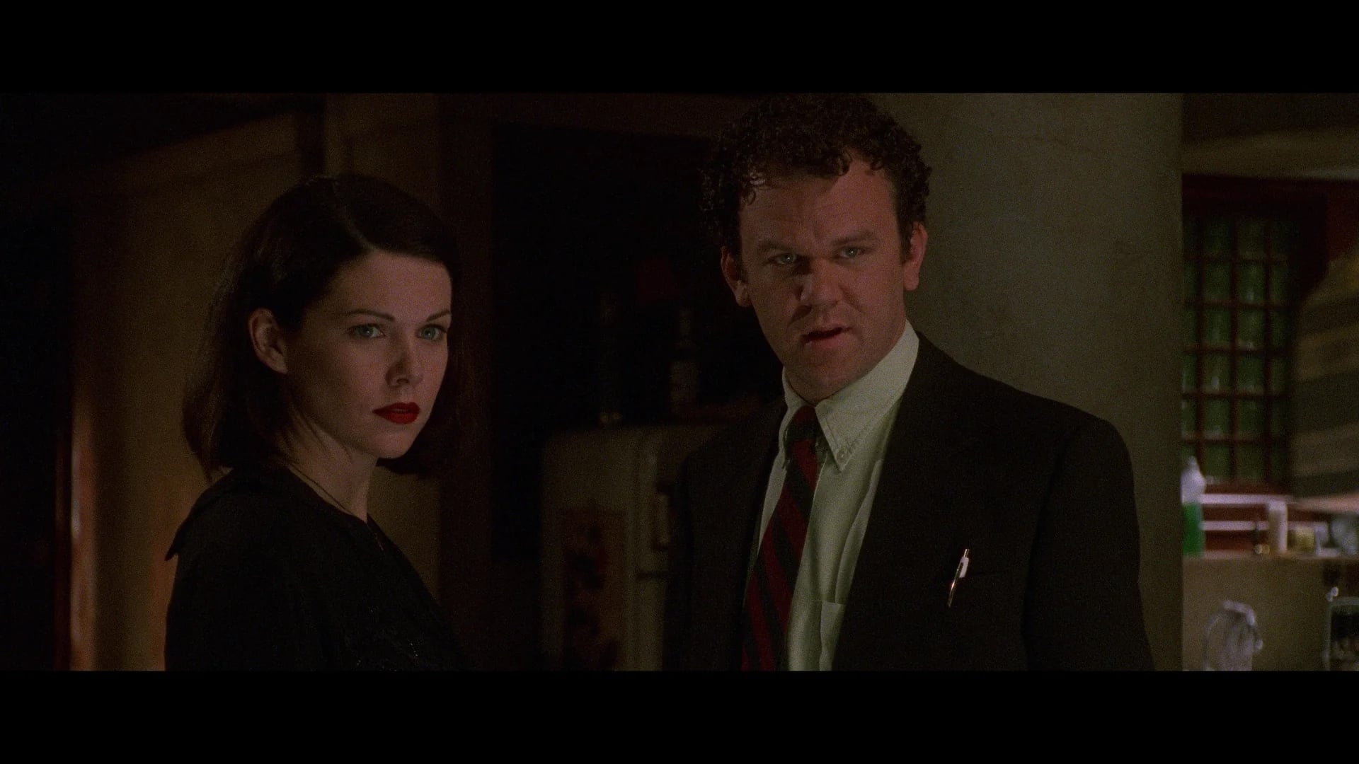 A woman with dark hair and red lipstick stands next to a man in a suit and tie inside a dimly lit room, both looking intently at something off-camera.