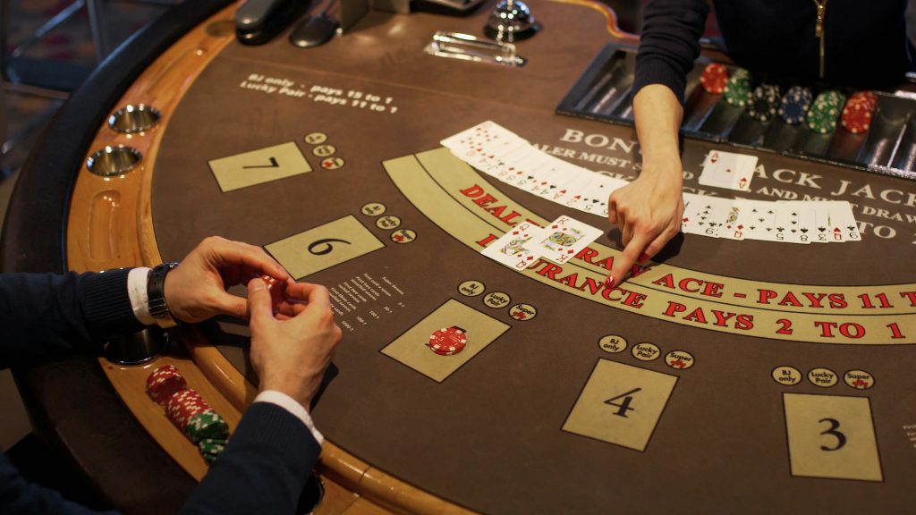 A person plays blackjack at a casino table, holding cards while a dealer displays two face-up cards. Chips are stacked on the table.
