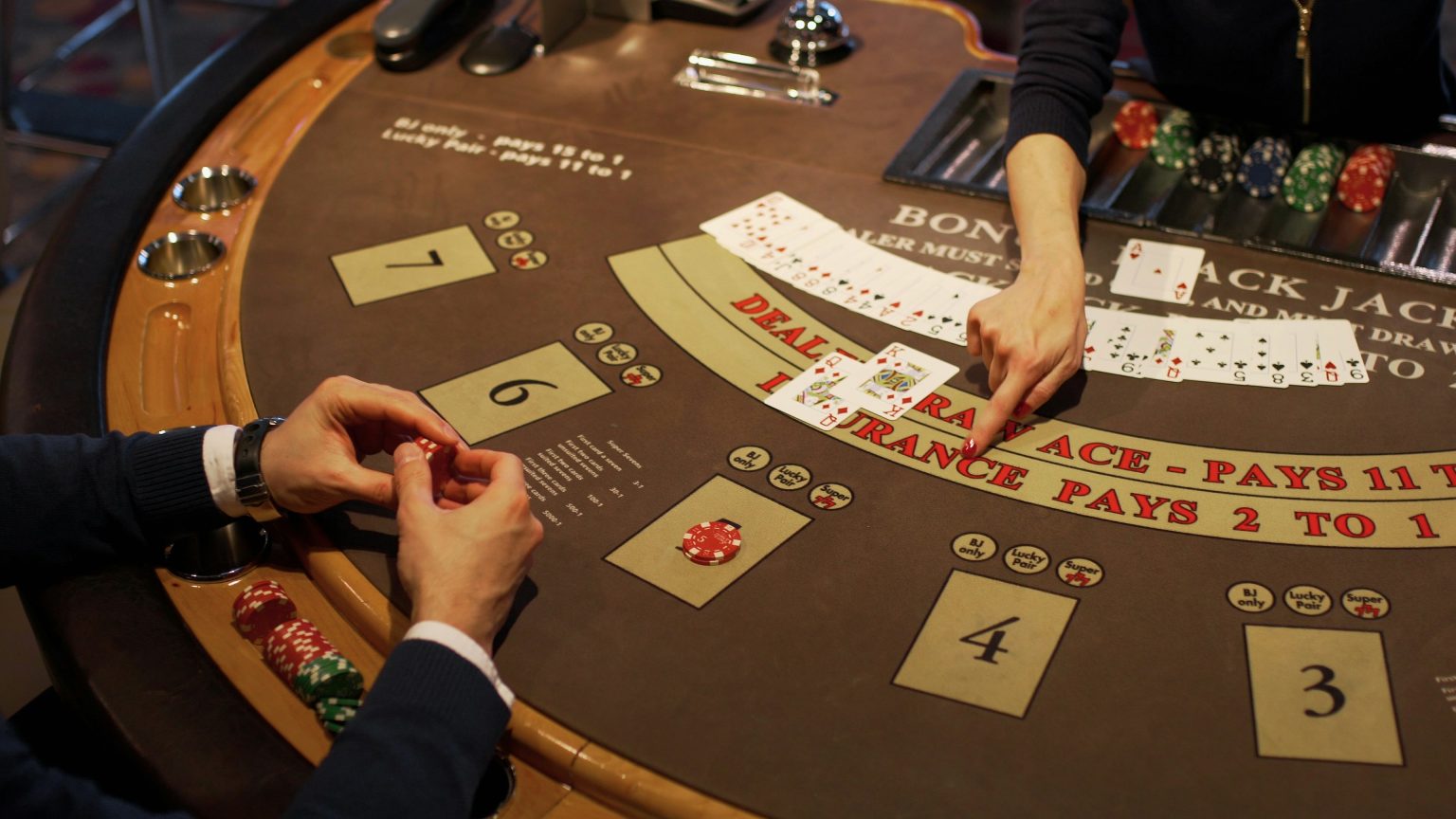 A person plays blackjack at a casino table, holding cards while a dealer displays two face-up cards. Chips are stacked on the table.