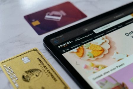 A tablet displaying the Amazon website sits on a white surface next to a gold American Express card and a partially visible purple card.