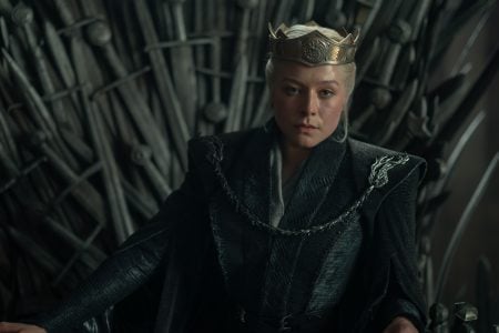 A woman wearing a crown and dark robes sits on a throne made of swords, looking forward with a serious expression.