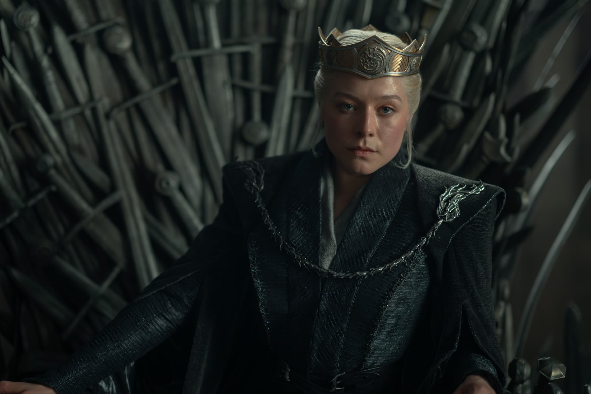 A woman wearing a crown and dark robes sits on a throne made of swords, looking forward with a serious expression.