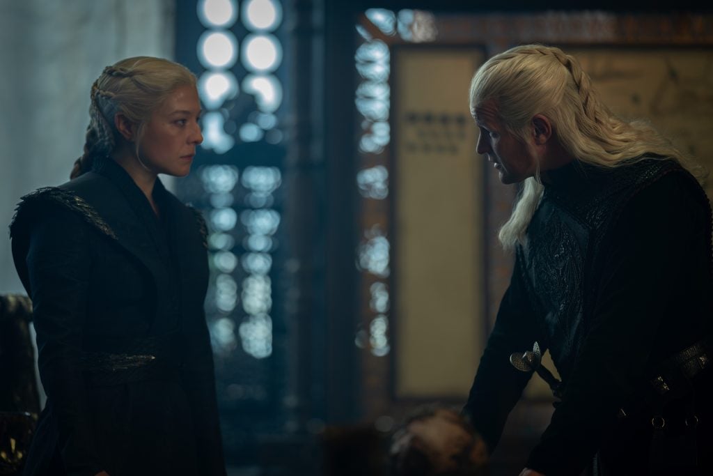 Two people with long, light hair and dark clothing face each other in a dimly lit room with ornate decor and patterned windows in the background.