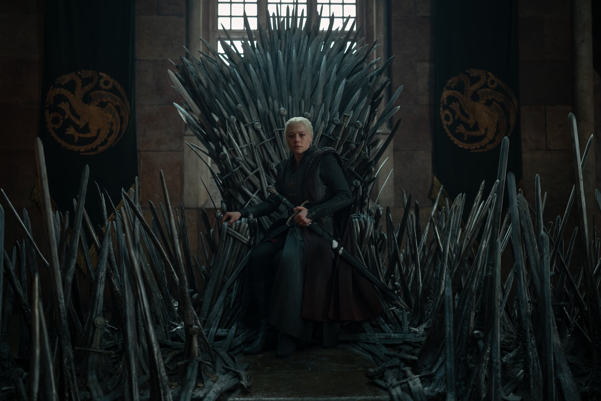 A person in medieval armor sits on an elaborate iron throne made of swords, with banners featuring dragon symbols on the walls behind.