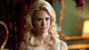 A woman with long, wavy blonde hair and a serious expression stands indoors in a softly lit room with a blurred background.