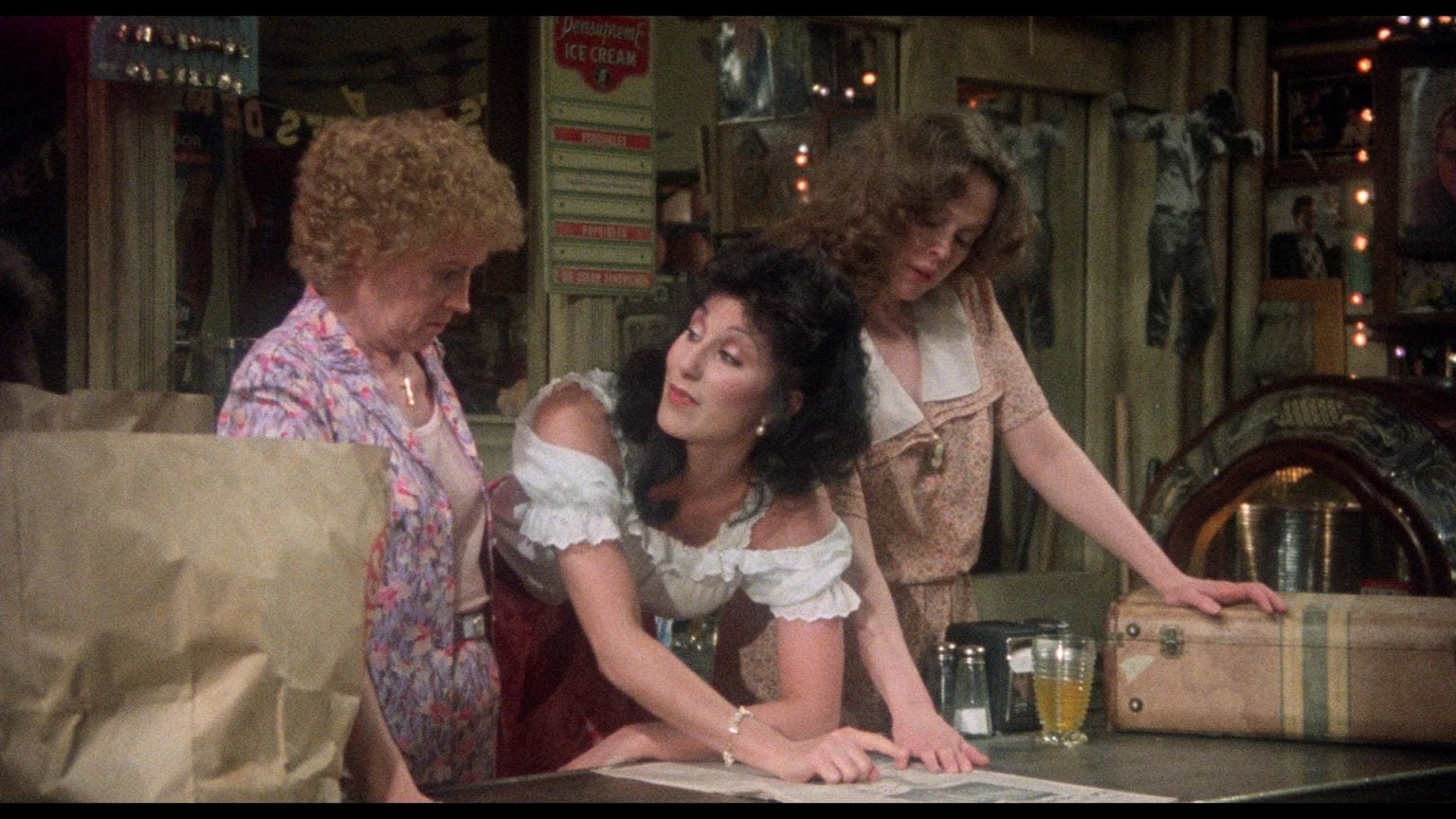 Three women stand around a counter in a vintage-style store, looking down at a newspaper spread out before them.