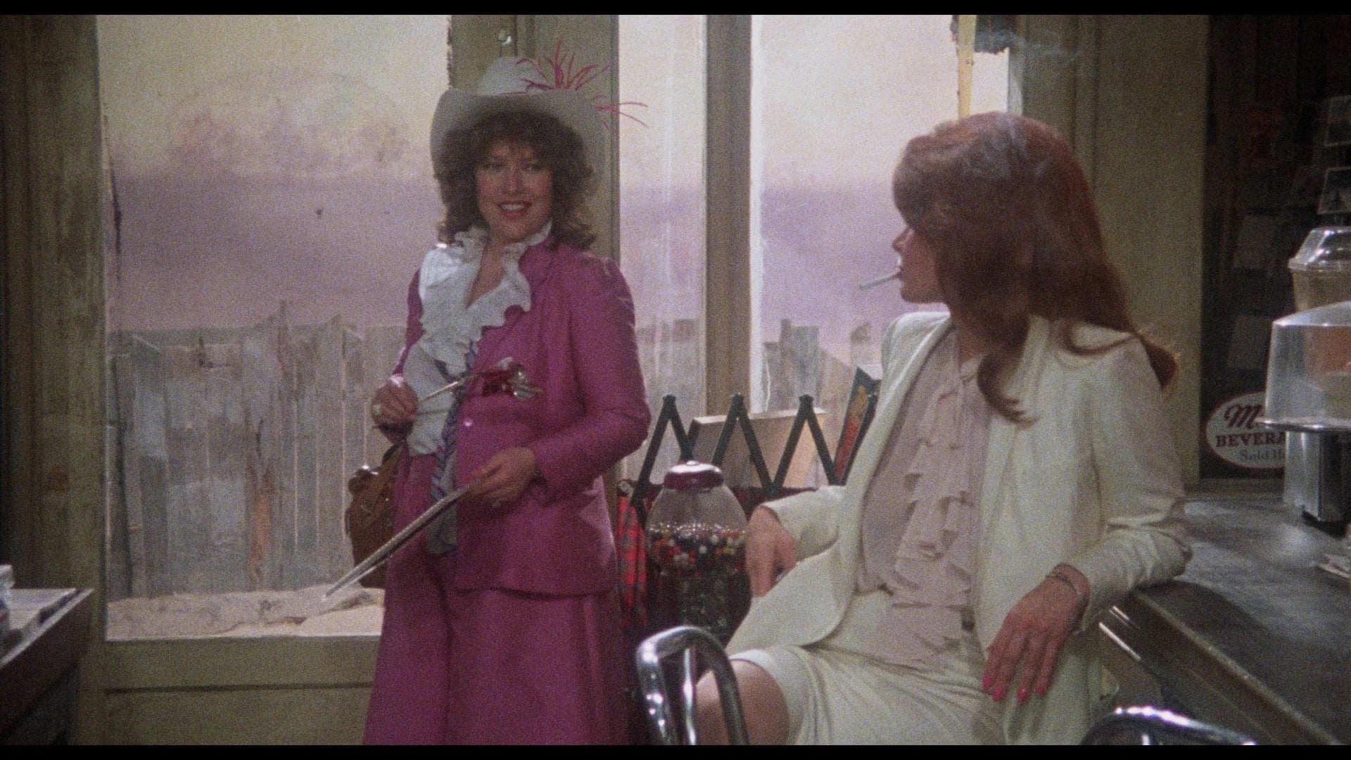 Two women talk in a diner; one stands in a bright pink outfit with a hat and cane, while the other sits at the counter in a white suit, near a gumball machine and a drink dispenser.