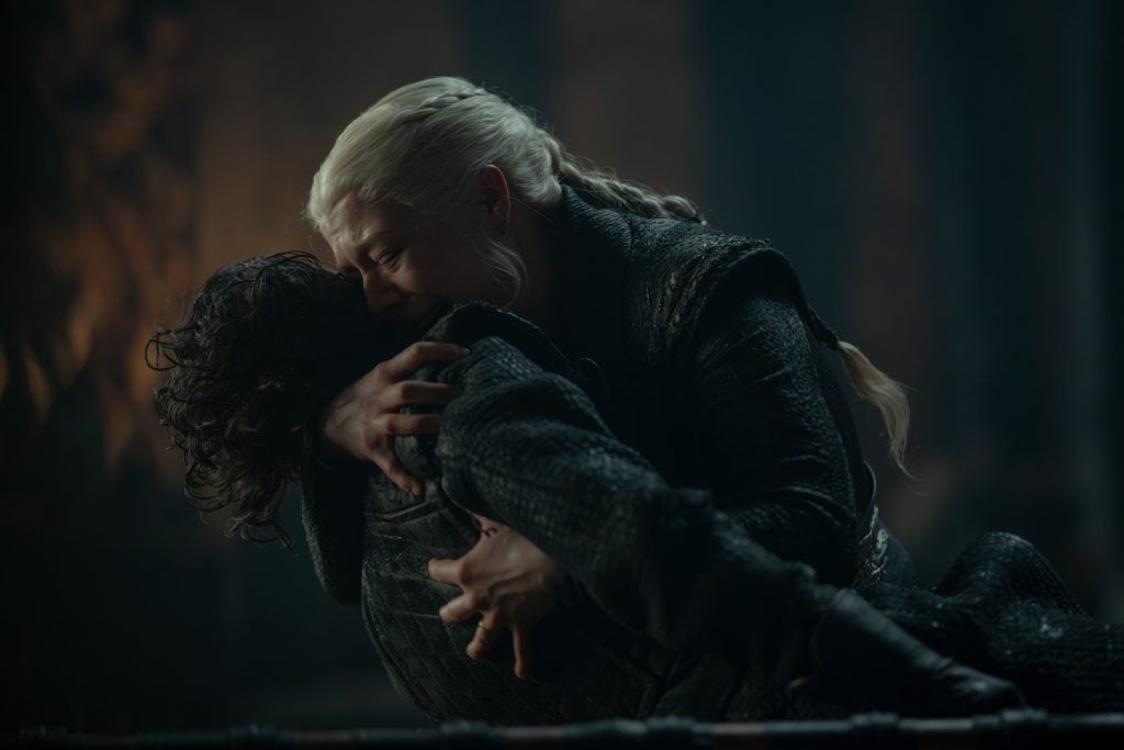 A woman with long, braided blonde hair embraces and comforts a person in dark clothing, both appearing emotional in a dimly lit setting.