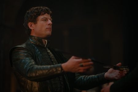 ‘House of the Dragon’ Season 3 Episode 4 Review: Ormund Becomes A Very Interesting Character A man in dark, ornate medieval attire stands indoors with his arms extended, holding a slender object.
