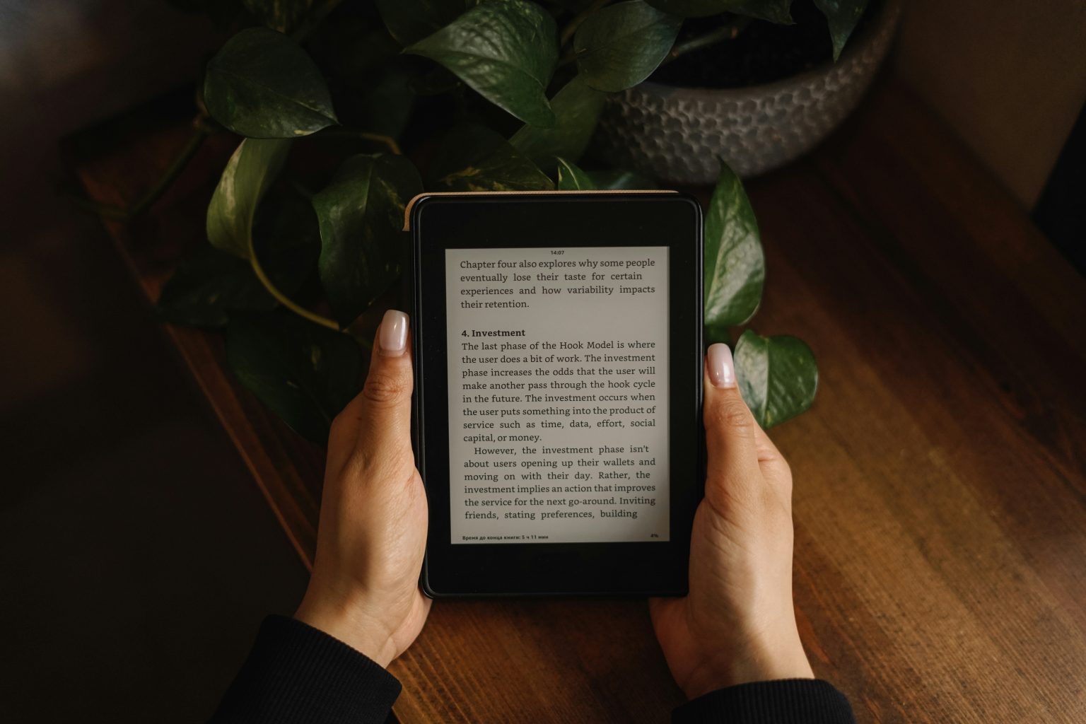 How To Convert PDF To AZW3 For Better Kindle Reading? Person holding an e-reader displaying text about investment, with a green plant and wooden surface in the background.